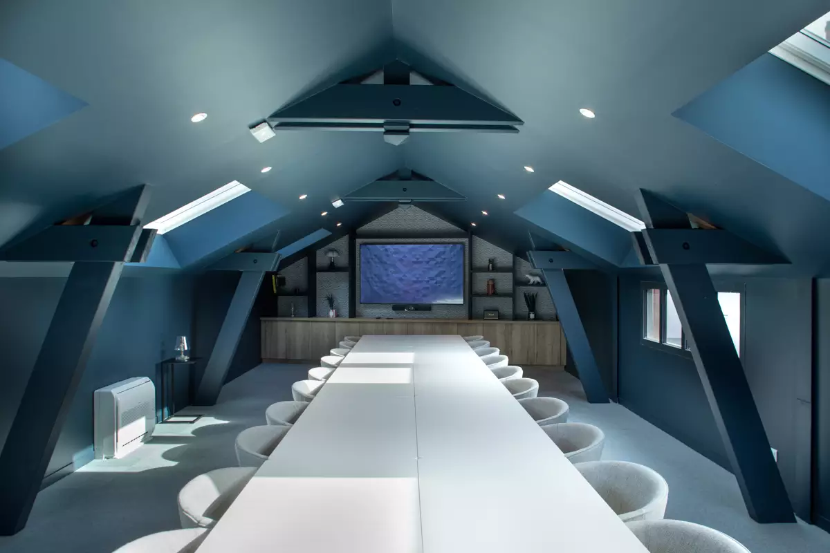 Salle de réunion avec plafond voûté, murs bleus, grande table blanche, chaises grises et écran près d'une étagère bois.