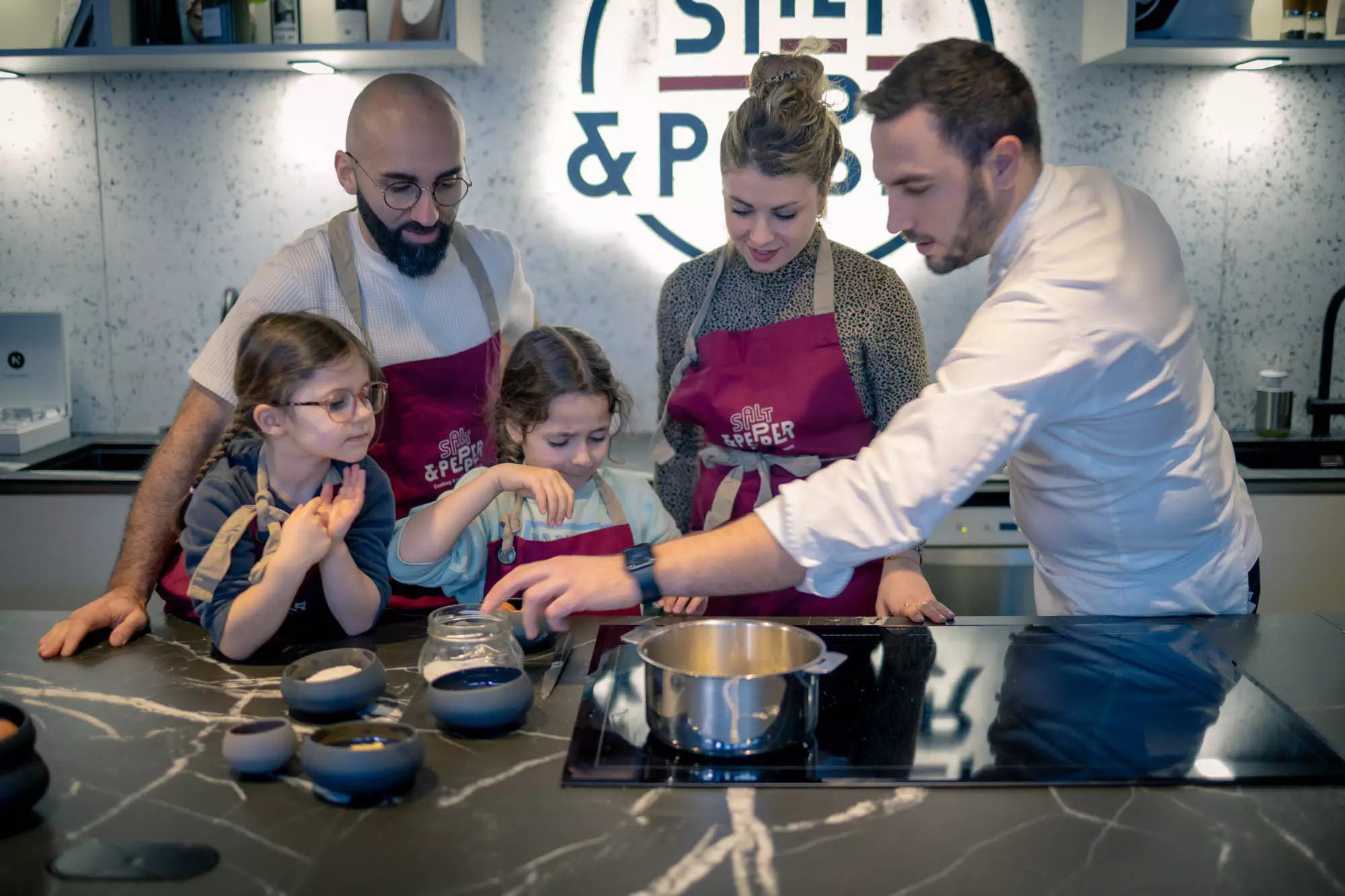 Chef teaching a cooking workshop to two adults and two children in aprons in front of an induction hob at Salt & Pepper Dijon.