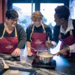 Trois personnes en tablier cuisinent ensemble et échangent autour d'un atelier culinaire dans une cuisine moderne à Dijon.