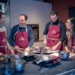 Groupe en tabliers rouges autour d'un îlot, préparant un plat ensemble chez Salt & Pepper Dijon, ustensiles à portée.