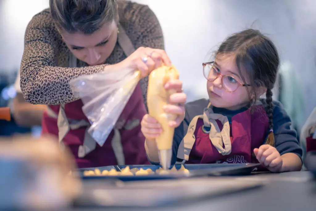Cours de pâtisserie : une femme guide une jeune participante à poche à douille sur une plaque, toutes deux en tablier.