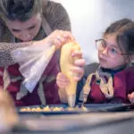 Cours de pâtisserie : une femme guide une jeune participante à poche à douille sur une plaque, toutes deux en tablier.