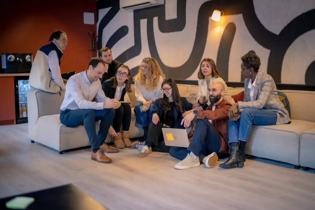 Groupe réuni autour d'un canapé, échanges animés lors d'un séminaire, certains avec ordinateur ou carnet.