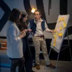 Animation team building : un homme présente des idées au paperboard devant deux collègues, décor design et lumière douce.