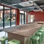Cuisine moderne avec ilot en marbre noir, tabourets verts, murs rouges et grandes fenêtres, idéale pour atelier culinaire.