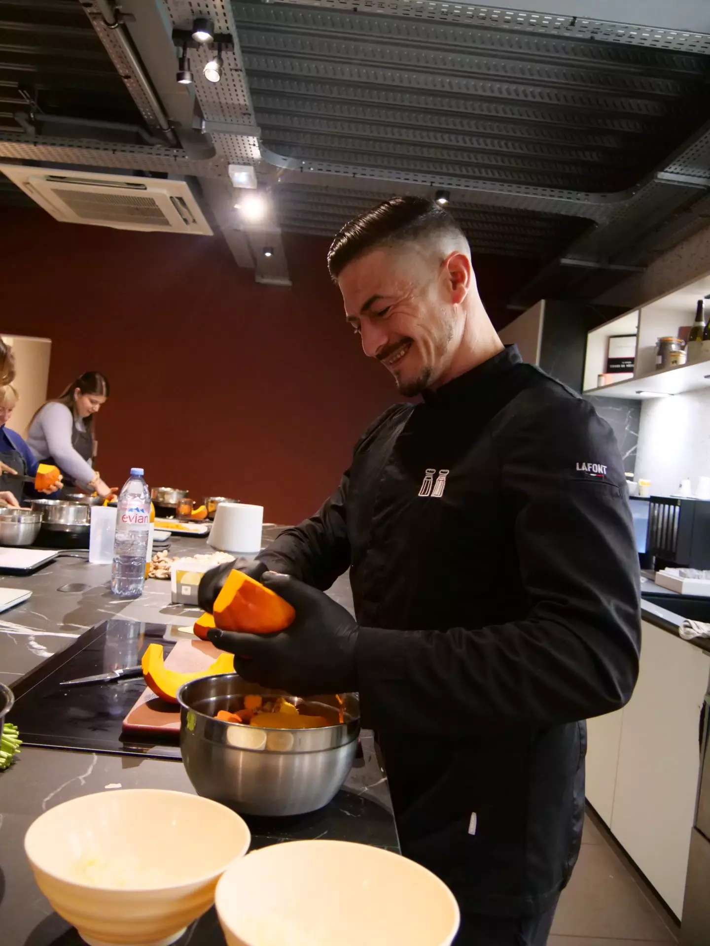 Homme en veste de chef noire et gants cuisine en équipe, tenant une courge orange au-dessus d'un bol chez Salt & Pepper Dijon.