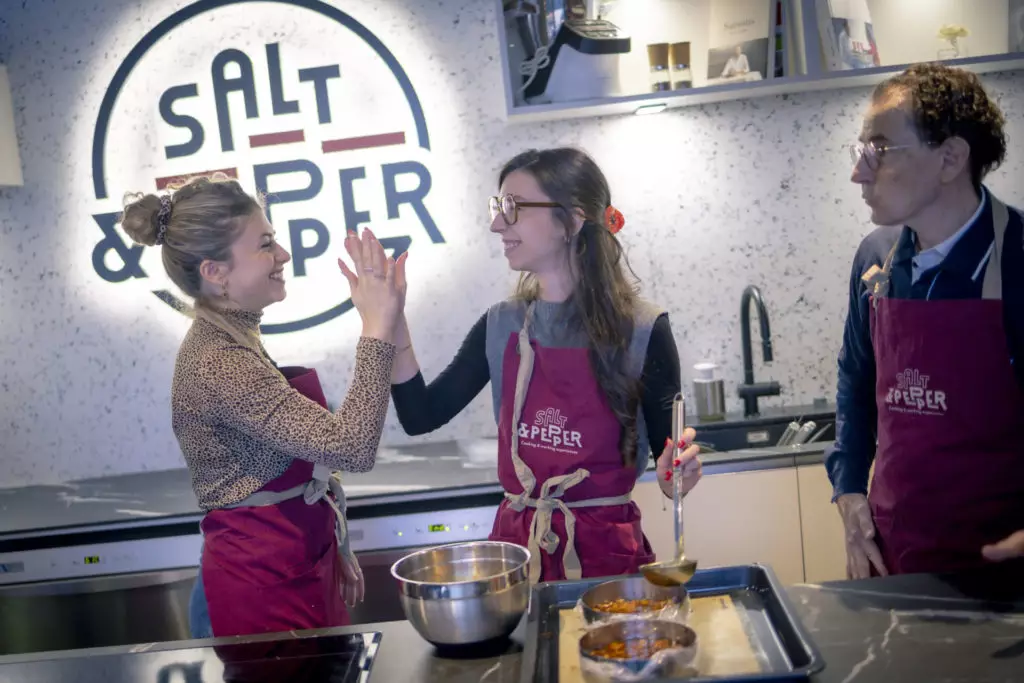 Deux femmes en tablier rouge se félicitent en cuisine, sous le regard d'un homme, pendant un atelier chez Salt & Pepper Dijon.
