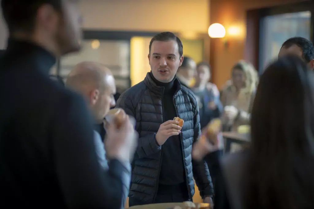 Un homme vêtu d'une veste noire tient une collation tout en discutant avec d'autres personnes dans un atelier culinaire à l'éclairage tamisé. Plusieurs personnes sont visibles à l'arrière-plan, ce qui suggère une réunion sociale décontractée ou un événement entreprise.
