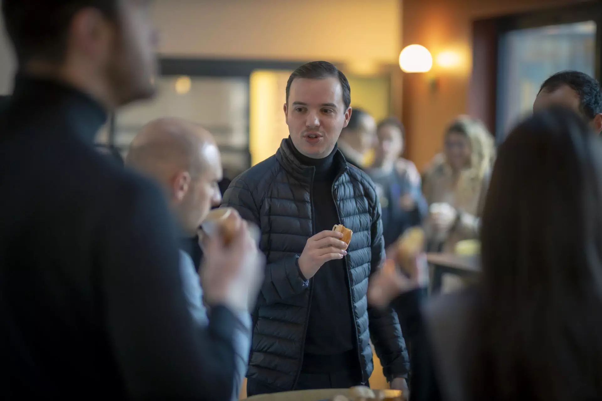 A man in a black jacket holds a snack while talking with other people in a dimly lit culinary workshop. Several people are visible in the background, suggesting a casual social gathering or a corporate event.