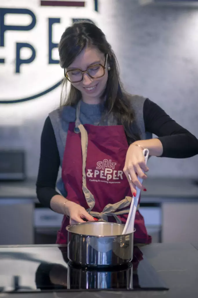 Une femme portant des lunettes et un tablier rouge Salt & Pepper sourit tout en remuant des aliments dans une casserole sur une cuisinière, dans une cuisine moderne.