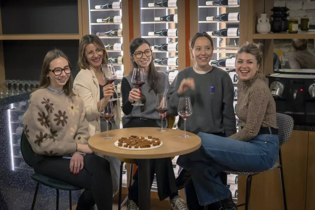 Cinq femmes sont assises autour d'une petite table avec des verres à vin et une assiette de snacks. Elles sourient et s'amusent dans un bar à vin moderne où les bouteilles sont exposées derrière elles.
